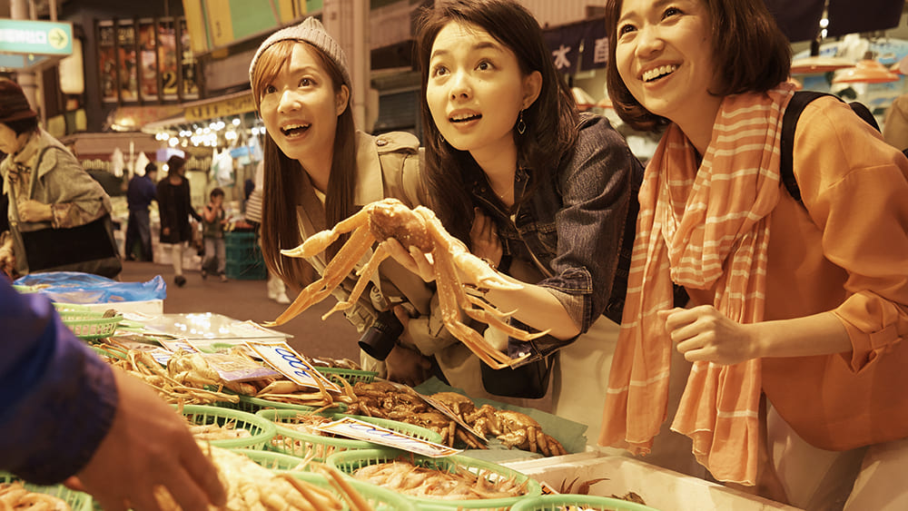市場に観光に来ている女性3人が店頭の商品を指さしている写真