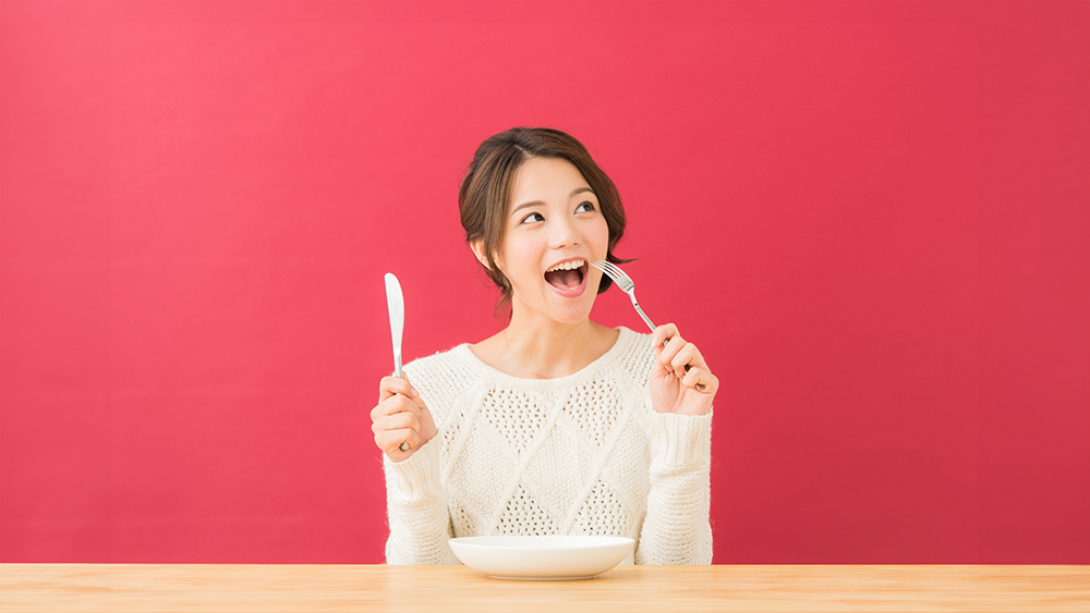 女性がナイフとフォークを持ち、食べる真似をしている写真