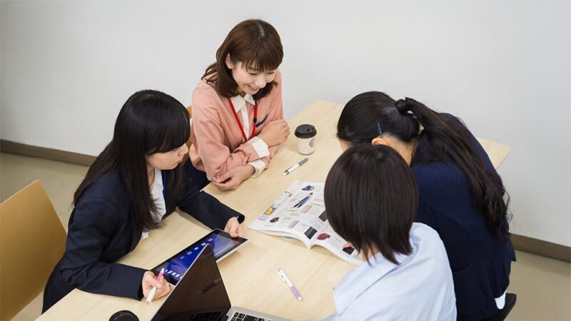スーツの女性4人が雑誌やパソコンを見ながら話し合っている写真