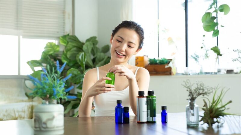 植物がたくさんある部屋の中で、笑顔の女性が緑のボトルを持って開けようとしている写真