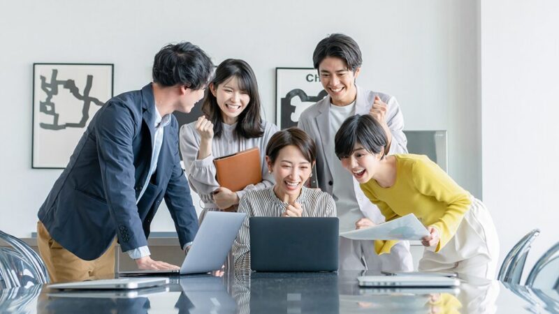 オフィスカジュアルの男女5人がパソコンを見て喜んでいる写真
