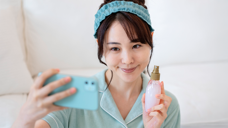 ヘアバンドをした女性が化粧品を持ってスマートフォンで自撮りしている写真