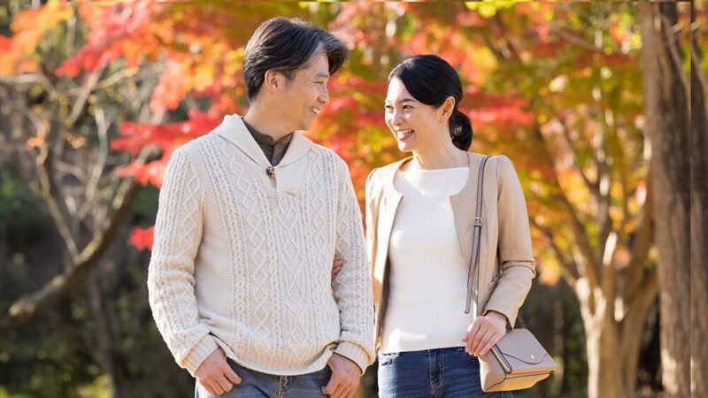ミドル世代の男女が紅葉の中を、笑顔で見つめあって歩いている写真