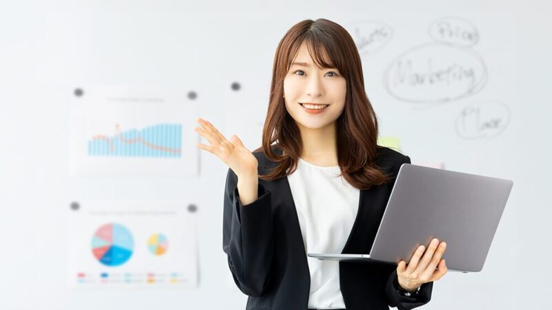 左手にパソコンを持ったスーツの女性が説明している写真
