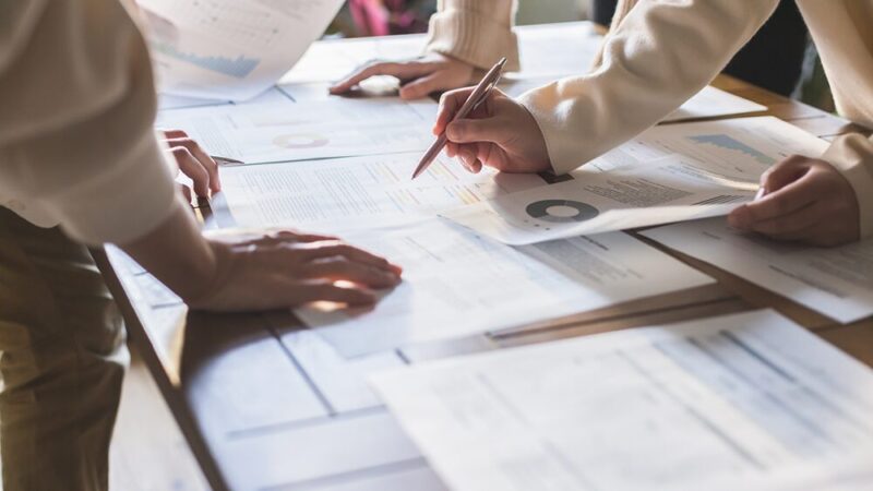 デスク上に並べられたたくさんの資料とそれを囲む社員の写真