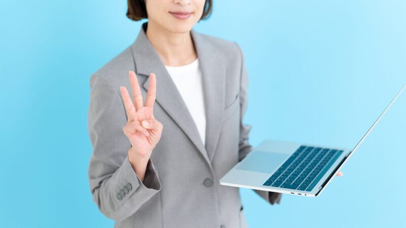 左手にパソコンを持ち、右手で3を示している女性の写真