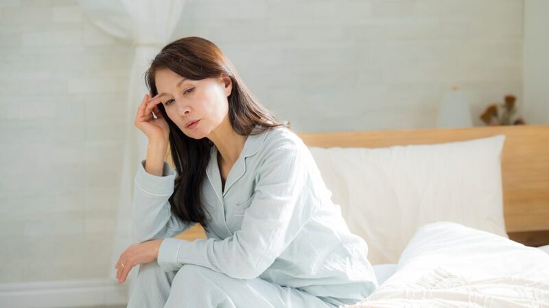 パジャマでベッド端に座り、暗い表情でこめかみに手を当てている中年女性の写真