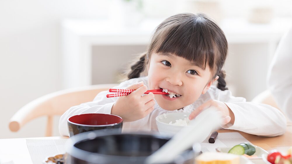 小さな女の子が箸で白飯を笑顔で食べている写真