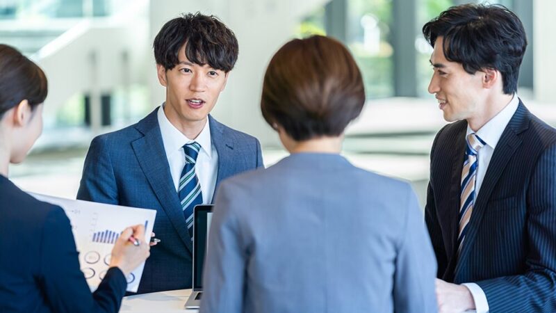 会議で資料を見ている女性とパソコンを見せて話している男性の写真