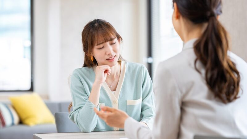顎に手を当てて悩んでいる女性と説明する女性の写真